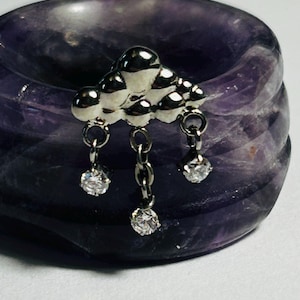 May include: A silver cloud charm with three hanging clear gemstones. The charm is set against a dark purple, textured background.