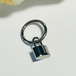 May include: A silver hinged ring with a small, rectangular padlock charm. The padlock has a rounded top and a small loop for attachment. The ring is circular and has a smooth, metallic finish. The background is a light, neutral colour.