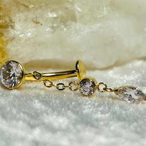 Puede incluir: Joyería de cuerpo dorada con una cadena y detalles de cristal transparente. La joyería tiene un cristal redondo en un extremo y un cristal en forma de lágrima en el otro extremo.