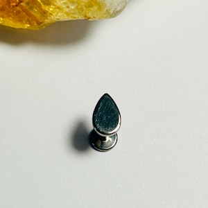 Può includere: Un orecchino a bottone a forma di goccia, color argento, con una base circolare piatta. L'orecchino è posto su uno sfondo bianco, con una pietra gialla e bianca nell'angolo in alto a sinistra.