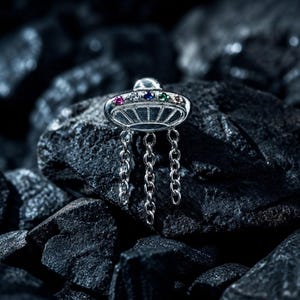 May include: A silver-toned UFO-shaped charm featuring five small, colorful inset gemstones along the top edge. The metallic saucer design includes three dangling silver chains hanging from the base, creating a distinct, multi-layered appearance.