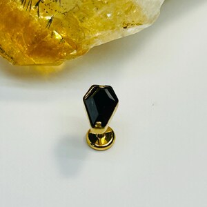 May include: A gold-toned body jewelry stud featuring a black, coffin-shaped gemstone set in a metal frame. The piece includes a flat, circular base and a post, designed for piercing, with a polished finish that reflects light on its surface.