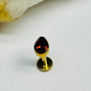 May include: A gold-toned flat back stud earring featuring a pear-shaped, deep red gemstone set in a bezel setting. The jewelry piece has a circular base plate and a short post, designed for body piercing, showcasing a polished metallic finish.