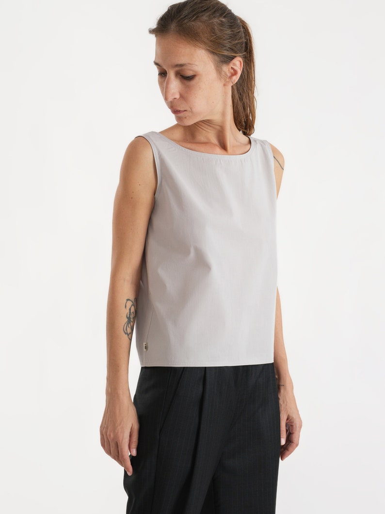 May include: A sleeveless, light grey, scoop neck top with a zip detail on the side. The top is cropped and has a relaxed fit.