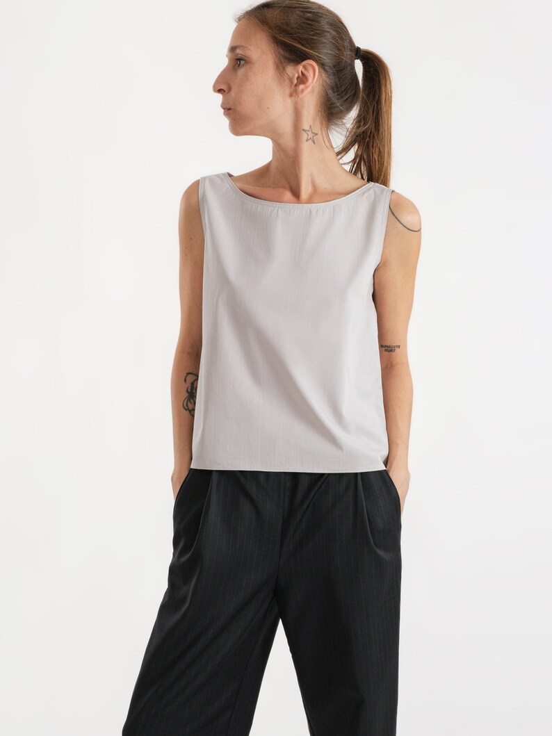 May include: A light grey sleeveless top with a boat neck and a relaxed fit. The top is paired with black trousers with a subtle pinstripe pattern.