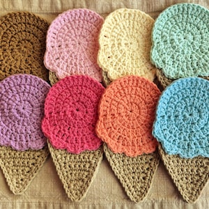 May include: A collection of eight handmade crochet ice cream cone coasters. Each coaster features a different color of ice cream, including brown, pink, yellow, green, purple, red, orange, and blue, atop a tan cone.