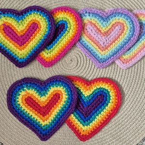 May include: A collection of handmade crochet heart-shaped coasters in various rainbow color schemes. Each coaster features a vibrant gradient of colors, including red, orange, yellow, green, blue, and purple, with a solid border. These coasters are perfect for adding a touch of color to any home.