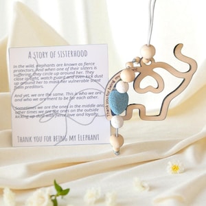 May include: Wooden elephant-shaped ornament with a string, featuring wooden and blue beads. The ornament has the text "Thank you for being my elephant" and a card with text about sisterhood.