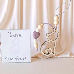 May include: A cat-shaped wooden ornament with a string, decorated with wooden beads and a heart-shaped bead. A card with the text "You're Puur-fect!!!" and a cat illustration is next to the ornament.