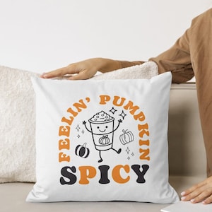 Könnte beinhalten: Ein weißes quadratisches Dekokissen mit einer zentralen Grafik einer lächelnden Kaffeetasse mit Armen und Beinen, umgeben von kleinen Kürbisillustrationen und Sternchen. Der Text lautet Feelin' Pumpkin Spicy in orangefarbener und schwarzer Schrift.