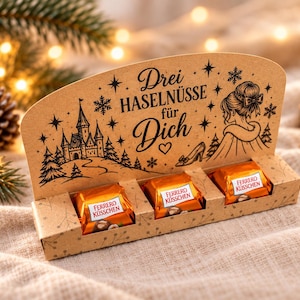Könnte beinhalten: Eine rechteckige Geschenkschachtel aus Karton mit drei Ferrero Küsschen Pralinen in orangefarbenen Verpackungen. Die Schachtel zeigt schwarze Druckgrafiken von einem Schloss, einer Person, einem Schuh, Schneeflocken und dem Text Drei HASELNÜSSE für Dich.