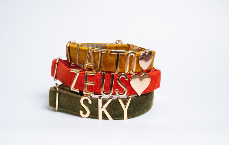 May include: Three velvet pet collars in mustard yellow, burnt orange, and olive green. Each collar features gold-toned metal letters and a heart charm. The top collar displays the text LATTO, the middle collar displays ZEUS, and the bottom collar displays SKY.