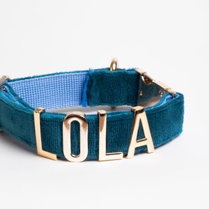 May include: A teal velvet pet collar featuring gold-toned metal letters that spell out the name LOLA. The collar includes a gold-colored metal buckle and a D-ring, with a light blue woven fabric lining visible on the interior side of the band.