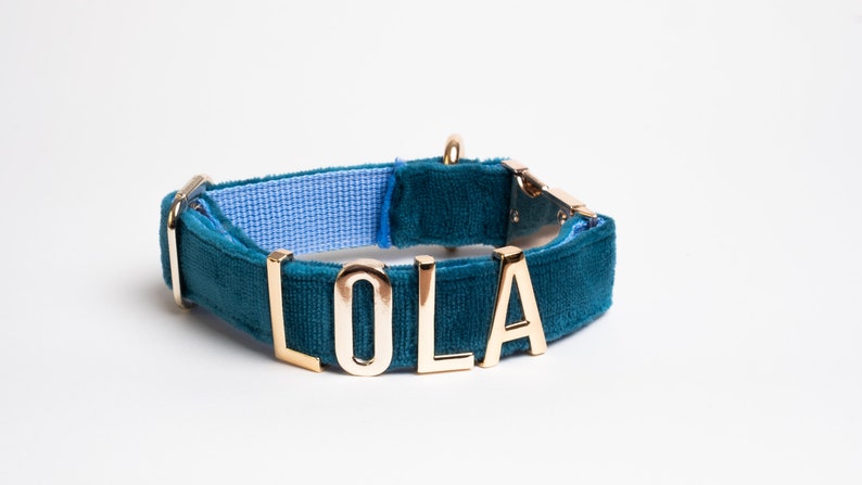May include: A teal velvet pet collar featuring gold-toned metal letters that spell out the name LOLA. The collar includes a gold-colored metal buckle and a D-ring, with a light blue woven fabric lining visible on the interior side of the band.