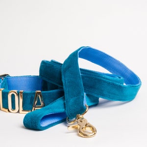 May include: A teal velvet pet collar and matching leash set featuring a bright blue interior lining. The collar displays the name LOLA in gold-toned metal letters. Both items include gold-toned metal hardware, including a swivel snap hook.