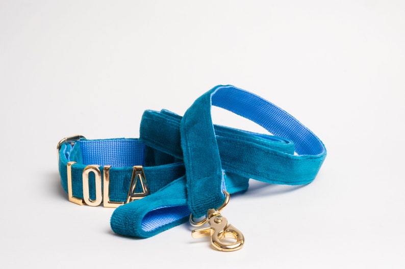 May include: A teal velvet pet collar and matching leash set featuring a bright blue interior lining. The collar displays the name LOLA in gold-toned metal letters. Both items include gold-toned metal hardware, including a swivel snap hook.