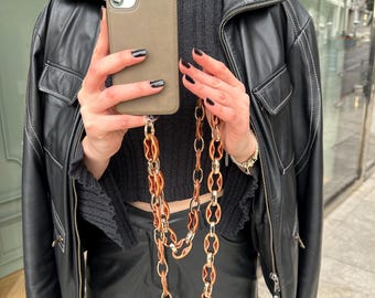 May include: A brown leather jacket with a brown and gold chain phone strap. The phone case is a muted green color.