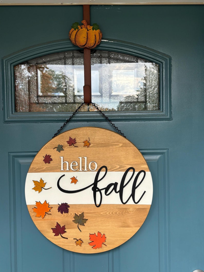May include: Round wooden fall door hanger with the words 'hello fall' in black script lettering on a white stripe.  The wood is light brown and features several small, layered autumn leaves in orange, red, and brown. The hanger has a black chain and is shown hanging on a teal door.