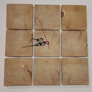 Puede incluir: Un reloj de pared de madera hecho de nueve piezas cuadradas de madera con una aguja de minutos roja y una aguja de horas negra. La madera tiene un patrón de veta natural.