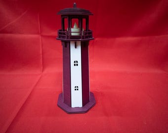 May include: A wooden lighthouse model with a burgundy exterior and white central panel. The lighthouse features small window cutouts and a lantern at the top. The model is set against a red background.