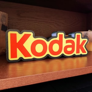 Könnte beinhalten: Ein schwarzes und gelbes Schild mit dem Wort "Kodak" in roten Buchstaben.