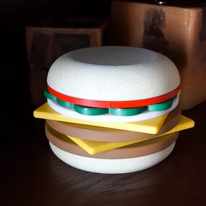 Puede incluir: Una hamburguesa de juguete blanca, gris, marrón, amarilla y verde con una franja roja y verde alrededor del pan superior.