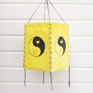 May include: A yellow paper lantern with a black and white yin yang symbol printed on it. The lantern has a fringe trim and is hanging from a string.