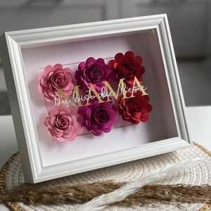 Könnte beinhalten: Weiß gerahmtes Blumenkunstwerk mit Papierblumen in Rosa- und Rottönen. Der goldene Text lautet "Mama Du bist die Beste". Das Kunstwerk wird auf einem gewebten Platzset präsentiert.