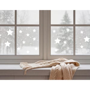 Könnte beinhalten: Eine winterliche Szene mit einem weißen Fenster mit sternförmigen Aufklebern. Ein cremefarbener Strickschal liegt auf der Fensterbank. Das Fenster blickt auf eine verschneite Landschaft mit Bäumen.
