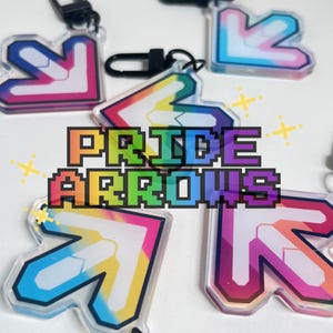 May include: A collection of acrylic keychains shaped like directional dance game arrows, featuring vibrant rainbow gradients and white centre accents. Each keychain includes a black lobster clasp. The text PRIDE ARROWS is overlaid in a pixelated font.