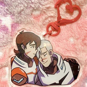 May include: A clear acrylic charm featuring two anime-style characters in a tender embrace. The charm has a heart-shaped clasp in a matching coral color. The characters are depicted in white, orange, and brown uniforms.