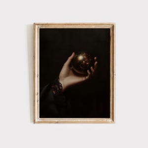 Peut inclure: Une main tenant un orbe rond foncé avec un motif doré tourbillonnant à l'intérieur. La main porte une manche en dentelle noire et l'arrière-plan est sombre.