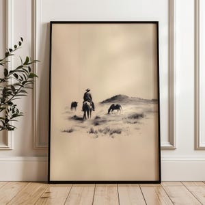 Könnte beinhalten: Ein gerahmter Sepia-Druck, der einen Cowboy auf einem Pferd zeigt, mit zwei weiteren Pferden und einem kleinen Hügel im Hintergrund. Das Kunstwerk ist im Bleistiftskizzenstil gehalten und hat einen schwarzen Rahmen. Die Szene erinnert an den Wilden Westen.