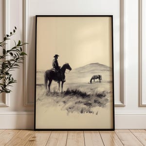 Könnte beinhalten: Gerahmter Schwarz-Weiß-Druck eines Cowboys auf einem Pferd in einem Feld. Das Kunstwerk zeigt einen Cowboy mit Hut, der ein Pferd reitet, mit einem anderen Pferd, das in der Ferne grast. Der Stil erinnert an eine Kohlezeichnung.