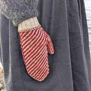 Könnte beinhalten: Ein rot-weiß gestreifter Fäustling wird in einem grauen Ärmel gehalten. Der Fäustling hat ein diagonales Streifenmuster und eine beige Manschette mit rot-weißem Zopfbesatz. Der Ärmel ist Teil eines grauen Pullovers. Der Hintergrund ist ein dunkelgrauer Stoff.