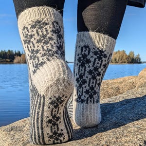 May include: A pair of knitted socks with a cream-colored base and dark gray geometric patterns. The socks have a ribbed cuff and a striped design on the foot. The socks are worn with black leggings, and the background shows a lake and blue sky.