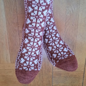 May include: Knitted socks in a warm brown hue, featuring a white floral design. The socks have ribbed cuffs, heels, and toes. The socks are displayed on a wooden surface.
