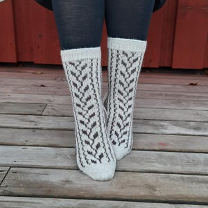 May include: A pair of mid-calf knit socks featuring a light cream base color with a dark brown vertical herringbone or leaf-like pattern running down the center. The socks have a ribbed cuff at the top and a solid cream toe section.