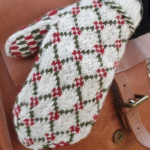May include: A knitted mitten in cream with a red and green geometric design. The cuff is cream with a green trim. The mitten is resting on a brown leather bag.