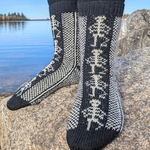 Puede incluir: Un par de calcetines estampados en blanco y negro con un diseño de árbol. Los calcetines son de punto y se llevan sobre leggings negros. El fondo presenta un lago y un cielo azul.