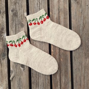 May include: A pair of beige knitted socks with a cherry pattern at the top. The cherries are red with green leaves. The socks are displayed on a weathered wooden surface.