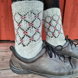 May include: A pair of gray argyle patterned socks with red and green accents, worn with brown leather shoes. The socks are knit with a diamond pattern. The shoes have black laces and a dark sole. The background is a red wooden surface.