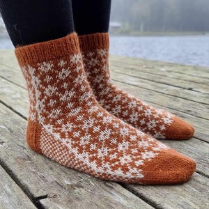 May include: A pair of knitted socks in shades of brown and white, featuring a geometric pattern. The socks have a ribbed cuff and a solid brown toe and heel. The socks are displayed on a wooden surface.