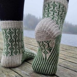 May include: Knitted socks featuring a green and cream tree design. The socks have a ribbed cuff and heel, with a striped pattern on the foot. The socks are displayed on a wooden dock, with a lake and trees visible in the background.