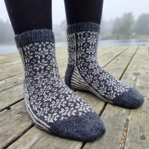 May include: A pair of knitted socks with a geometric pattern in shades of gray and white. The socks have a dark gray cuff, heel, and toe. The socks are worn on a wooden dock.