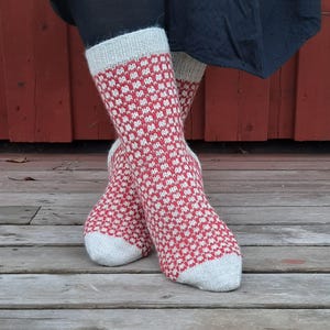 May include: A pair of red and white patterned socks. The socks have a repeating geometric design in red on a white background. The cuff and toe of the socks are white. The socks are worn on a wooden surface.