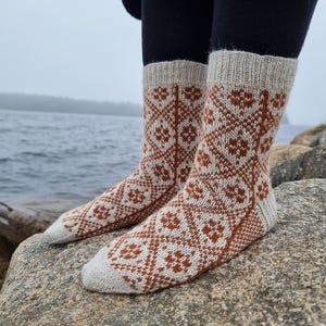 May include: A pair of patterned socks with a beige cuff, heel, and toe, and a brown and white geometric design. The socks are knit and appear to be made of wool. The socks are worn by a person standing on a rock near the water.