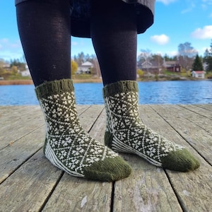 May include: A pair of green and white patterned socks. The socks have a geometric design and are worn with black leggings. The socks are on a wooden dock with a lake in the background.