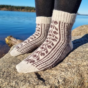 Puede incluir: Un par de calcetines de punto con estampado beige y marrón. Los calcetines presentan un diseño detallado con motivos geométricos y florales. Los calcetines se llevan con leggings negros y descansan sobre una roca cerca de una masa de agua.