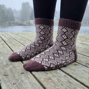 May include: A pair of knitted socks with a geometric pattern in shades of brown and cream. The socks have a ribbed cuff and heel, and are worn over black leggings. The socks are on a wooden surface.